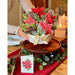 Decorative Christmas centerpiece with poinsettias and pinecones on a wooden tray, set on a table with candles and a card.
