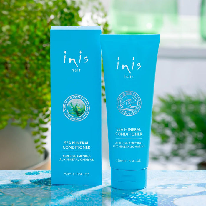 Blue Inis Hair Sea Mineral Conditioner tube (250ml / 8.5 fl. oz.) displayed upright on a wooden table with a soft-focus bathroom setting, starfish accent, and glass stones nearby.