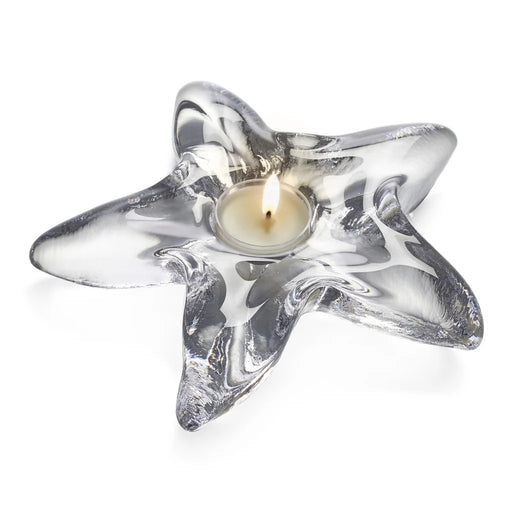 Clear glass candle holder containing a lit candle, casting a warm glow in a softly lit environment.