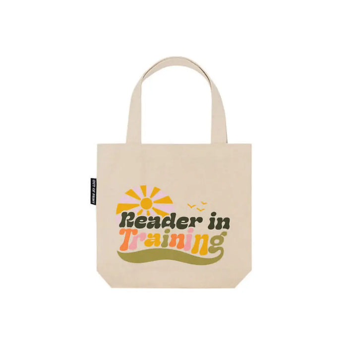 Tote bag with 'Reader in Training' text on a table next to a book in a white background 