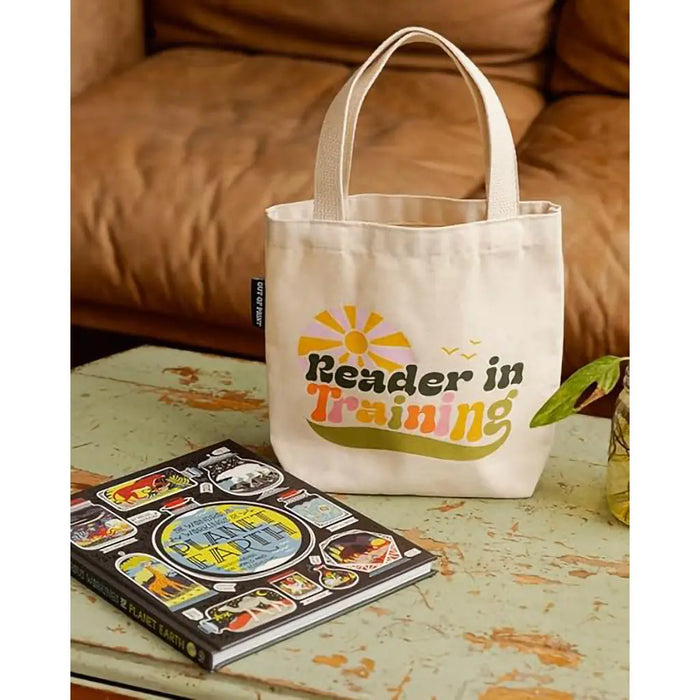 Tote bag with 'Reader in Training' text on a table next to a book
