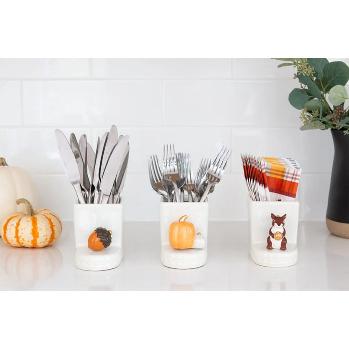 Three white containers with cutlery, decorative pumpkins, and a potted plant on a light surface.