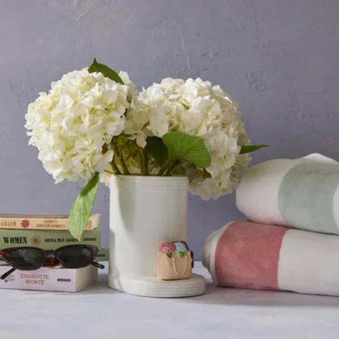 White flowers in a vase on books with sunglasses, a small Nora Fleming Handbag Mini , and folded towels against a gray background.