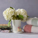 White flowers in a vase on books with sunglasses, a small Nora Fleming Handbag Mini , and folded towels against a gray background.