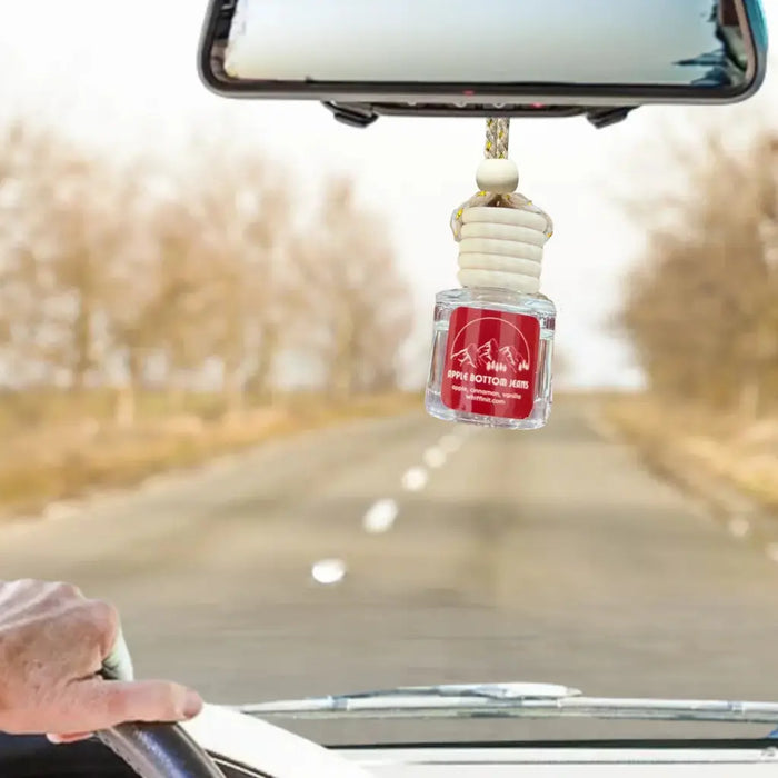 A car air freshener bottle labeled 'Apple Bottom Jeans' hanging from a car's rearview mirror while a hand is visible on the steering wheel, with a blurred road and trees in the background.
