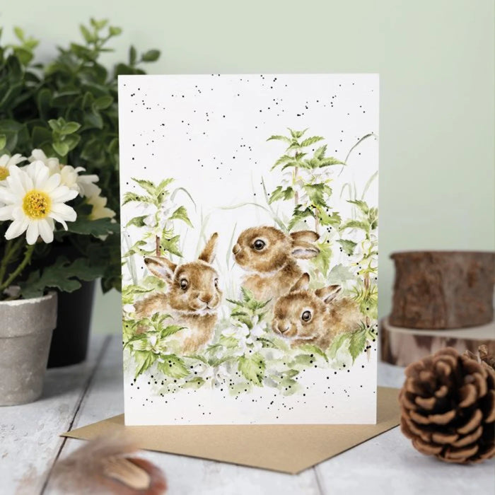 A greeting card depicting three rabbits frolicking in a grassy field, surrounded by nature.