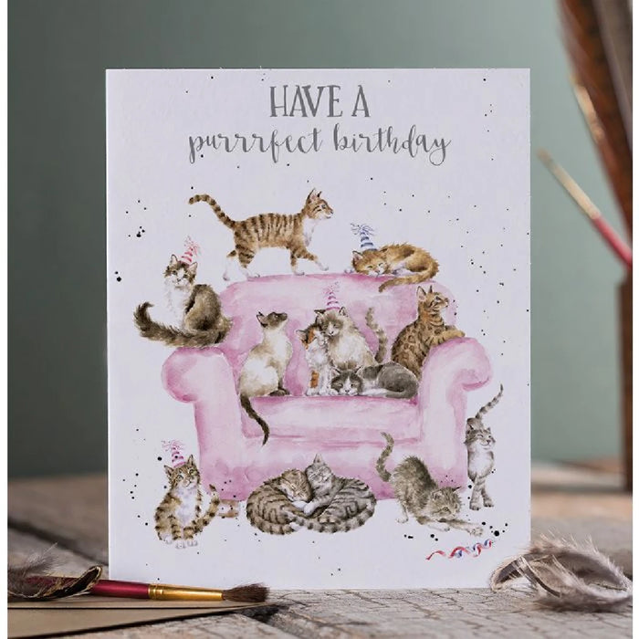 A whimsical birthday card showing cats on a pink couch, celebrating with vibrant colors and decorations.