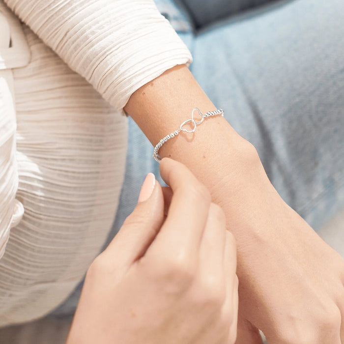 A Littles & Co. A Little Friendship bracelet worn on wrist featuring silver beaded stretch design and interlinked heart charm