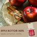 Apple Bottom Jeans diffuser bottle shown with a warm-toned background of red apples and cinnamon sticks on a wooden board, labeled 'Apple Bottom Jeans' with scent notes 'apple, cinnamon, vanilla'.