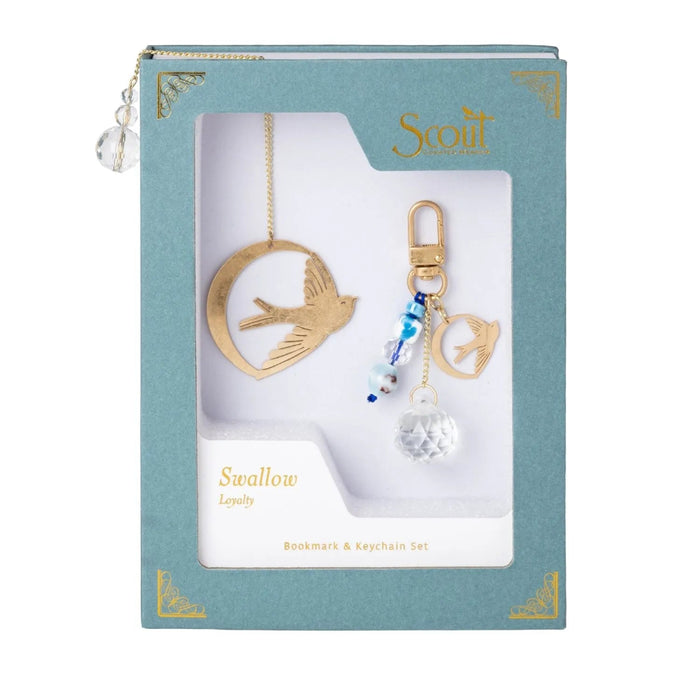 Scout Curated Wears Swallow/Loyalty bookmark and keychain set on a pale pink background. Gold-tone bookmark features a swallow with outstretched wings inside an open circular outline, attached to a long chain with a clear faceted charm