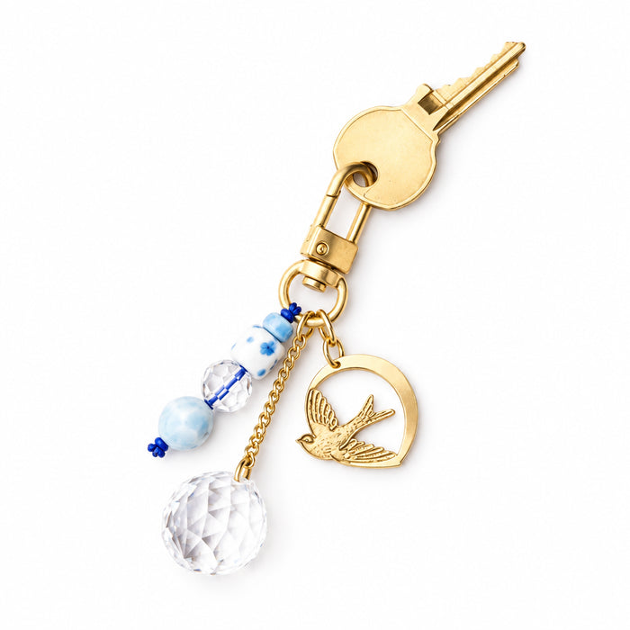 Scout Curated Wears Swallow/Loyalty bookmark and keychain set on a pale pink background. Gold-tone bookmark features a swallow with outstretched wings inside an open circular outline, attached to a long chain with a clear faceted charm