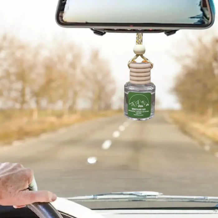 A car air freshener bottle hanging from a rearview mirror in a car, with a blurred road and trees in the background. The bottle has a green label that reads 'Mountain View Car Air Freshener'.