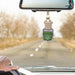 A car air freshener bottle hanging from a rearview mirror in a car, with a blurred road and trees in the background. The bottle has a green label that reads 'Mountain View Car Air Freshener'.