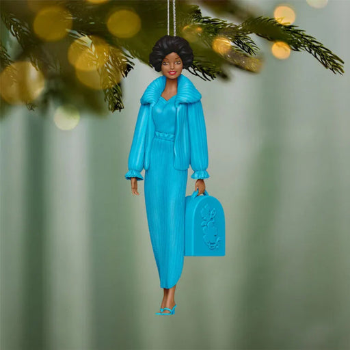 Ornament of a woman in a blue outfit holding a suitcase, hanging from a tree.