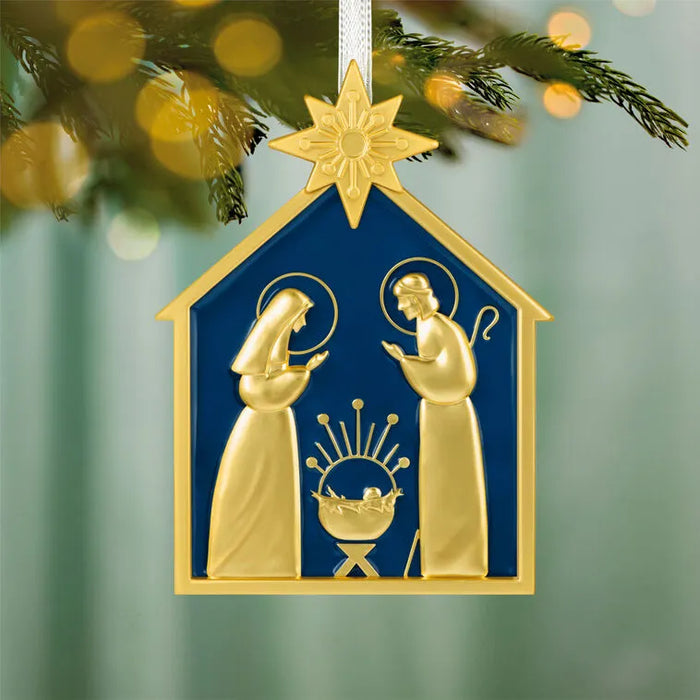 Close-up of Hallmark 2026 metal nativity ornament hanging from white ribbon on pine branch with bokeh lights