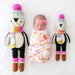 Newborn baby lying between two Cuddle + Kind Aspen the Penguin dolls, one large and one small, wearing colorful scarves and winter hats.