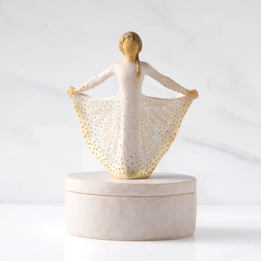 A decorative figurine of a ballerina with outstretched arms holding a skirt, displayed on a round marble-like box against a soft white marble background.