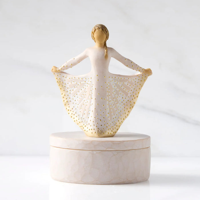 A decorative figurine of a ballerina with outstretched arms holding a skirt, displayed on a round marble-like box against a soft white marble background.