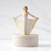 A decorative figurine of a ballerina with outstretched arms holding a skirt, displayed on a round marble-like box against a soft white marble background.