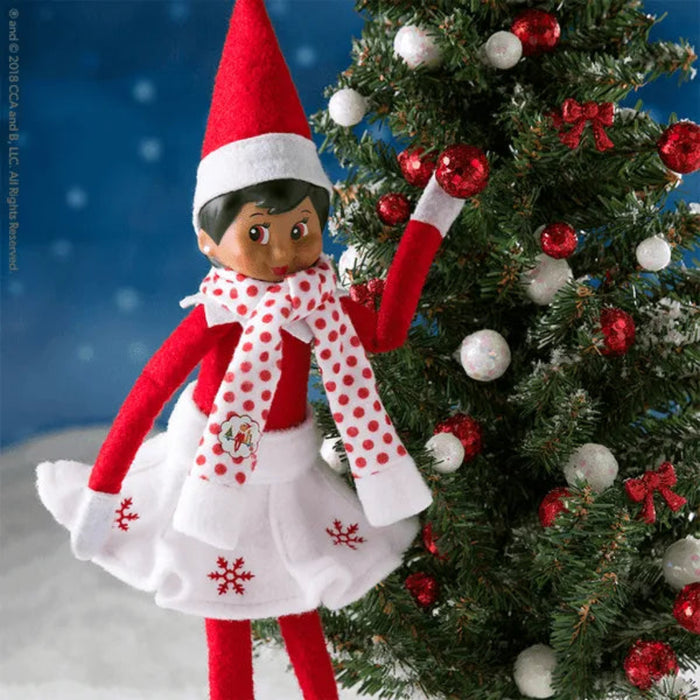 Elf on the Shelf in a red and white outfit standing next to a decorated Christmas tree.