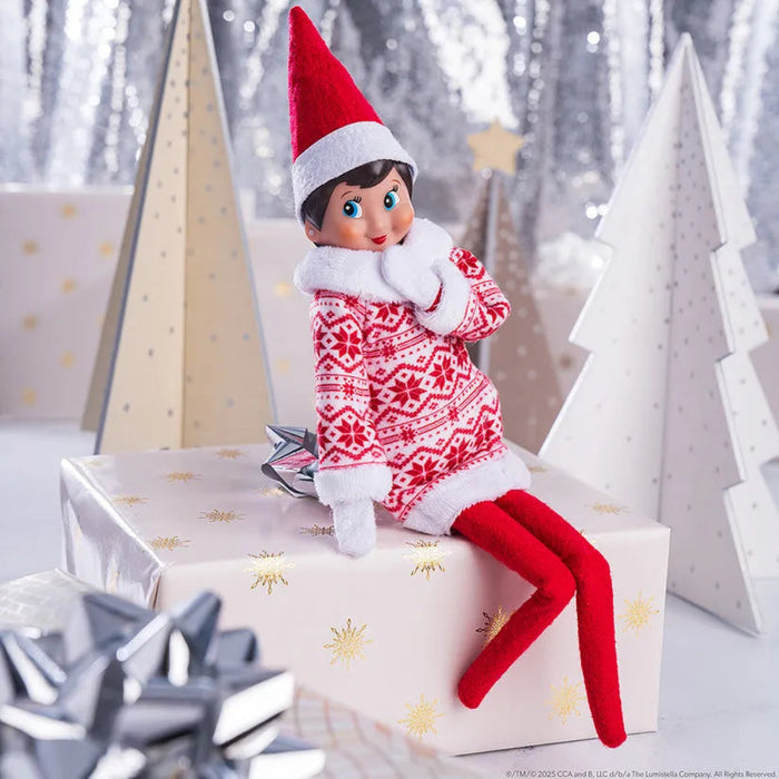 Elf on the Shelf in a red and white outfit sitting on a gift box with Christmas decorations in the background.