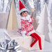 Elf on the Shelf in a red and white outfit sitting on a gift box with Christmas decorations in the background.