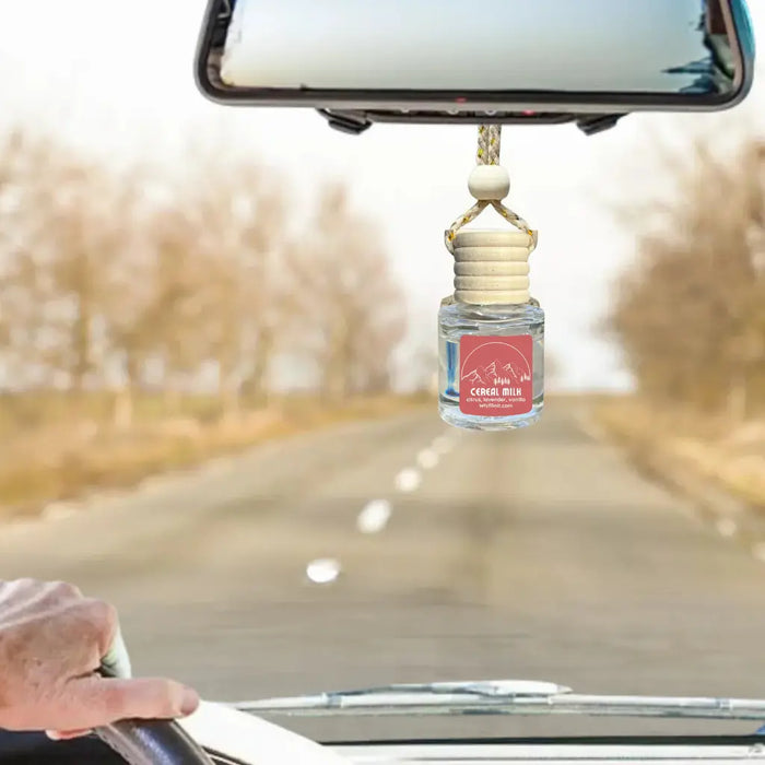 A car air freshener Cereal and Milk  bottle with a wooden dropper cap hangs from the rearview mirror inside a moving car. The bottle has a clear glass body with a red and white mountain logo that reads 'Coral Bisc'.