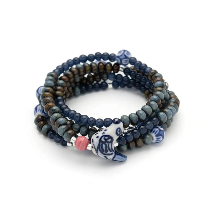 Scout Curated Wears Peace/Dove ceramic bead wrap bracelet/necklace with dark navy, muted green, and bronze-toned round beads, silver-toned spacers, blue-and-white patterned ceramic accents, and a white dove-shaped charm with blue detailing.