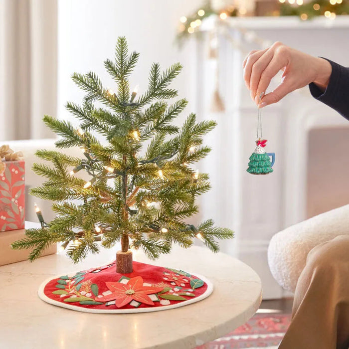 Green tree mug ornament hanging on lit Christmas tree with fireplace background display