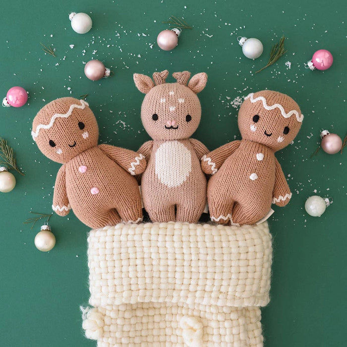 Three Cuddle + kind knitted toys resembling gingerbread men and a deer on a textured surface with Christmas ornaments in the background.