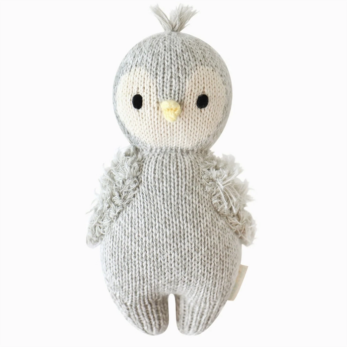 Front view of a hand-knit baby penguin doll in gray and cream yarn with a yellow beak, black stitched eyes, fuzzy wings, and a tuft of yarn on its head.