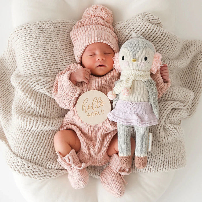 Newborn baby in a pink knit outfit and hat laying on a cream blanket, holding the little size Skylar the Penguin doll dressed in earmuffs, scarf, and lavender skirt.