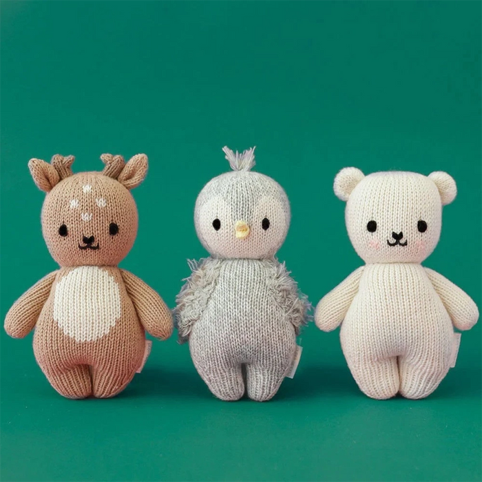 Three hand-knit baby dolls lined up against a green background: a fawn with antlers, a gray penguin with fuzzy wings, and a cream bear.