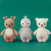 Three hand-knit baby dolls lined up against a green background: a fawn with antlers, a gray penguin with fuzzy wings, and a cream bear.