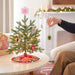 Small decorated Christmas tree on a table with a hand placing an  mini princess ornament, in a cozy living room setting.