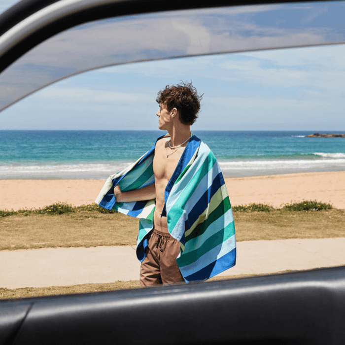 Man standing by the ocean wearing Dock & Bay Color Canyon striped quick dry towel in shades of green, blue, and yellow