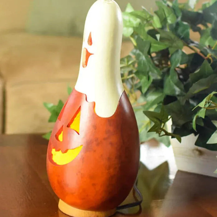 A carved gourd Halloween decoration lamp with a white ghost figure on top, glowing yellow eyes and mouth, and a wooden base, sitting on a wooden table with a blurred indoor background and green plant.