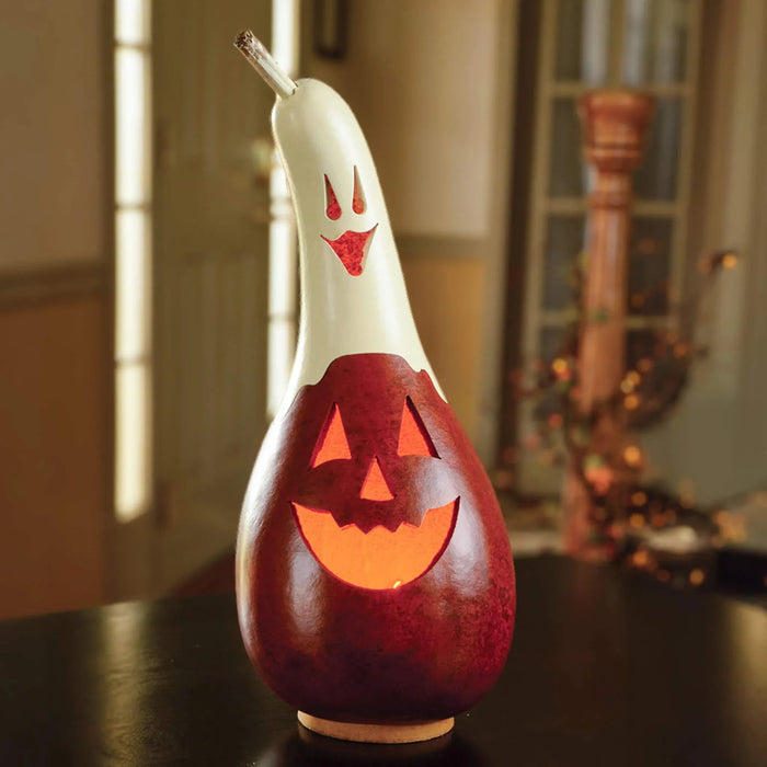 A carved gourd Halloween decoration with a white ghost face on top and a glowing orange jack-o'-lantern face on the body, set on a dark table in a softly blurred indoor room.