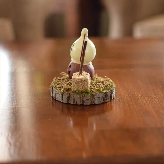 A small decorative figurine of a character sitting at a campfire on a rustic wood slice base, placed on a polished wooden table.