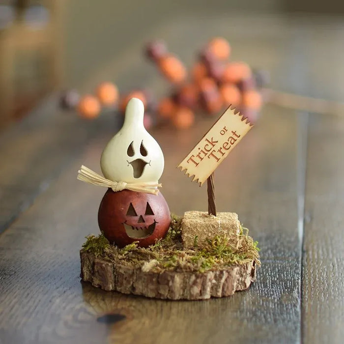 A small Halloween tabletop decoration set on a rustic wood slice base, featuring a carved brown gourd pumpkin, a white ghost figure, faux grass, moss, a small stone-like block, and a sign that reads 'Tuck or Treat.' The scene is photographed on a wooden table with blurred orange pumpkins in the background.
