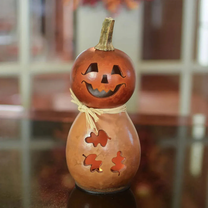 A carved natural gourd decoration shaped like a jack-o'-lantern with a smiling face, leaf-shaped cutouts on the body, and a small stem, photographed on a reflective surface with a softly blurred indoor background.