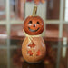 A carved natural gourd decoration shaped like a jack-o'-lantern with a smiling face, leaf-shaped cutouts on the body, and a small stem, photographed on a reflective surface with a softly blurred indoor background.