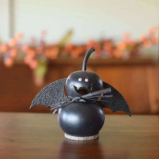 A black Halloween decorative figure shaped like a gourd with bat wings, a small twig stem, and a black bow around the neck, displayed on a wooden surface with a blurred indoor background.