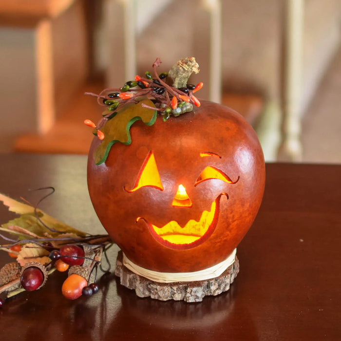 A carved orange jack-o'-lantern pumpkin candle with a glowing face, decorated with small autumn leaves, berries, and a twig on top, sitting on a wood slice base on a dark wooden table.