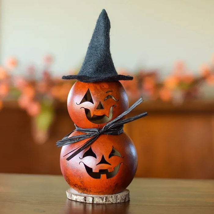 A decorative two-tiered gourd Halloween lantern with carved jack-o'-lantern faces, a black pointed witch hat, a black ribbon bow, and a small round wooden base, shown on a tabletop with a blurred indoor background.