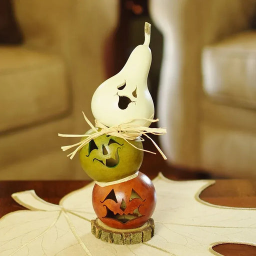 A stacked Halloween decoration made from three carved gourds: a white pear-shaped gourd on top, a green pear in the middle, and an orange pumpkin at the bottom, tied with raffia and displayed on a leaf-shaped mat in a softly blurred indoor setting.