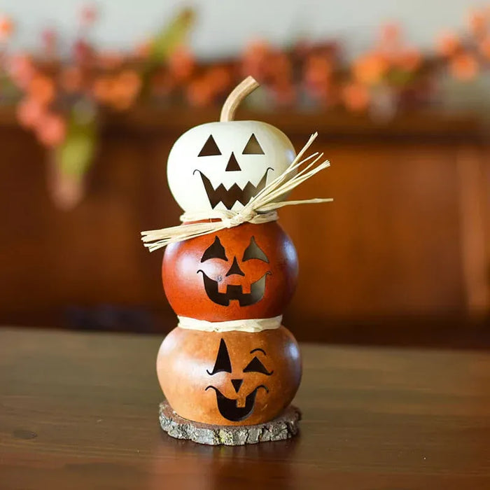A stacked Halloween decoration made from three carved gourds with jack-o'-lantern faces, tied together with dried raffia, displayed on a wooden surface with a blurred indoor background.