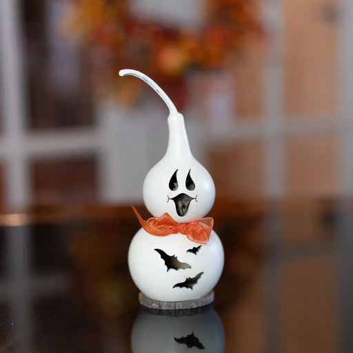 A white ceramic Halloween gourd decoration with a ghost face, an orange bow tie, and bat cutouts, displayed on a reflective dark surface with a blurred indoor background.