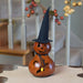A carved gourd Halloween decoration shaped like a witch's head, with a black pointed hat, black facial features, a small black twig scarf, and a glowing yellow candle inside, displayed on a tabletop with a blurred staircase background.