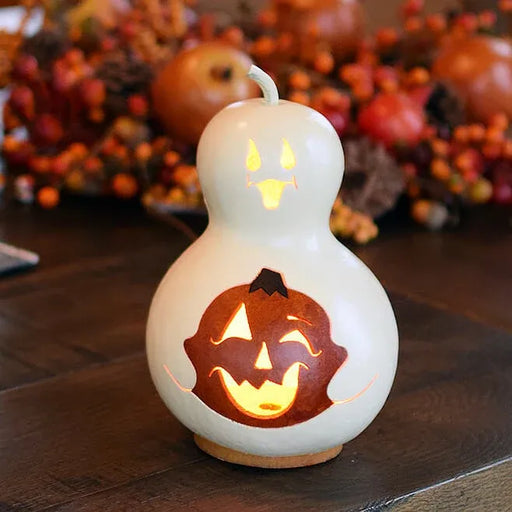 A white gourd-shaped candle with a carved jack-o'-lantern face, glowing with warm yellow light, set on a dark wooden table with autumn decorations blurred in the background.